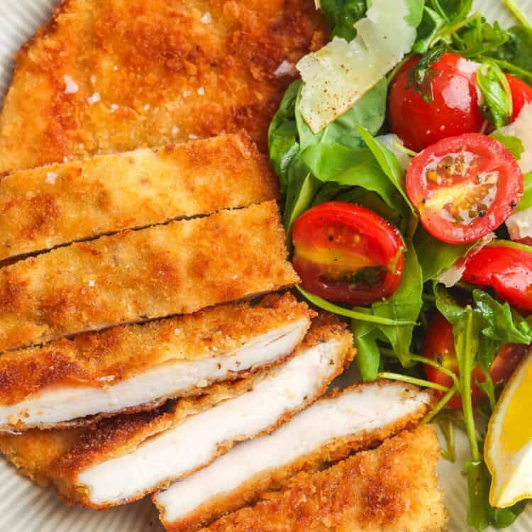 a dinner plate of sliced chicken milanese with a side of arugula salad with tomatoes.