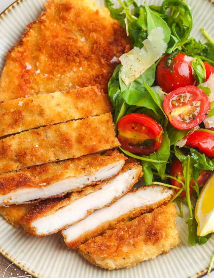 a dinner plate of sliced chicken milanese with a side of arugula salad with tomatoes.