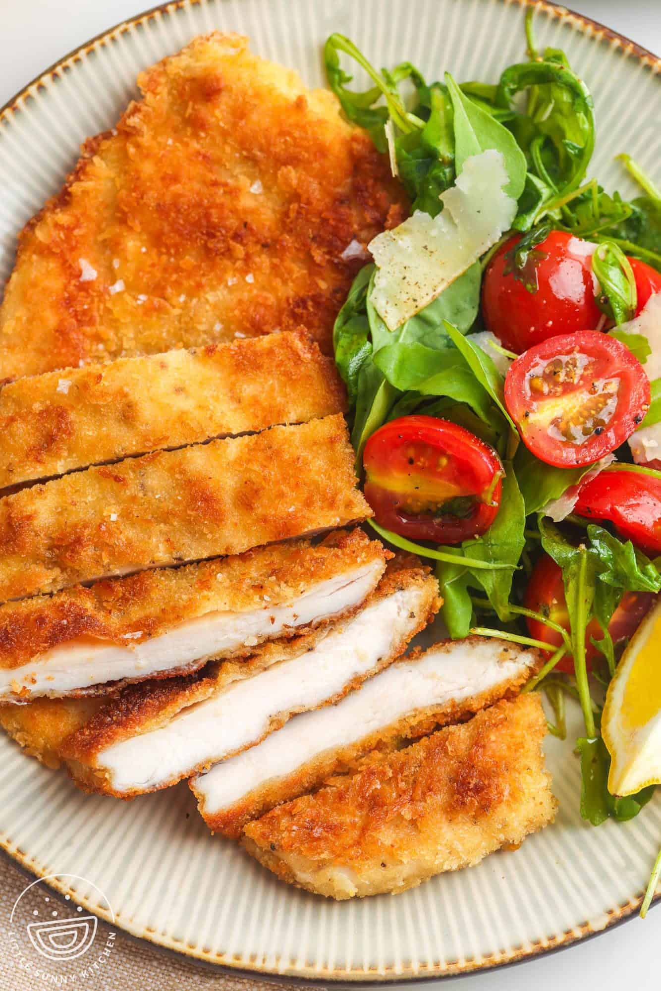 a dinner plate of sliced chicken milanese with a side of arugula salad with tomatoes.