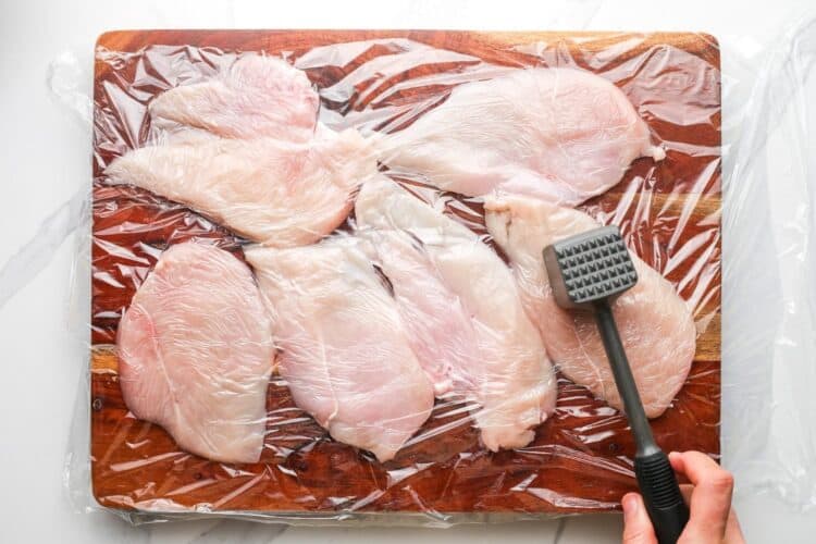 chicken cutlets on a wooden board, underneath plastic wrap. A hand is using a mallet to pound the chicken out into even, thin pieces.