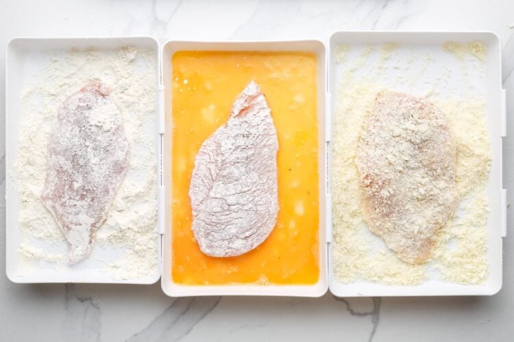 chicken cutlets being breaded in a three step system using rectangular plastic trays. The first tray is flour, the second is beaten egg, and the third is panko breadcrumbs. Each tray also has a chicken cutlet in it.