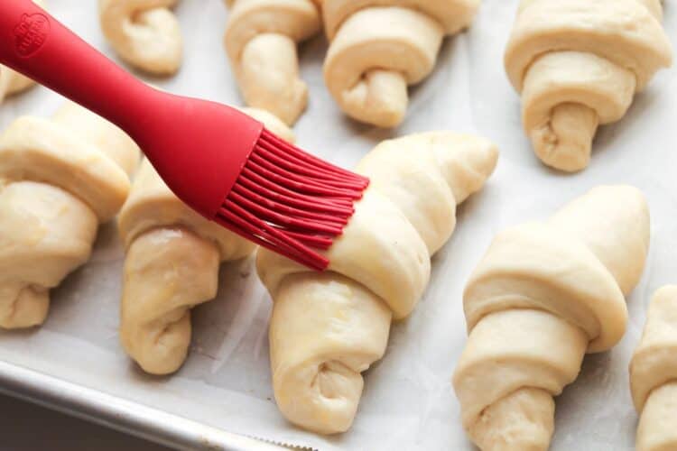 unbaked crescent rolls on a baking sheet. A red brush is adding egg wash to the tops of each.