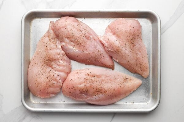 four chicken breasts on a small pan, seasoned with salt and pepper.