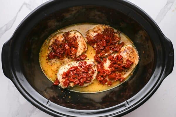 four chicken breasts added to a slow cooker with broth. The chicken is topped with chopped sun dried tomatoes.