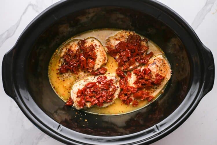 four chicken breasts added to a slow cooker with broth. The chicken is topped with chopped sun dried tomatoes.