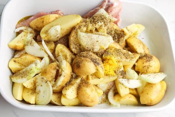 Chicken thighs mixed with quartered yellow potatoes and onions in a baking dish, all seasoned with lemon zest and dried herbs.