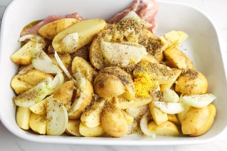 Chicken thighs mixed with quartered yellow potatoes and onions in a baking dish, all seasoned with lemon zest and dried herbs.