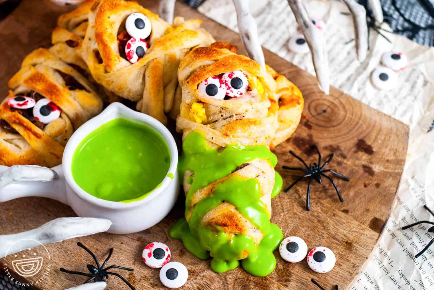 Halloween breakfast mummies made with pizza dough wrapped around eggs, sausage, and bacon, decorated with candy eyeballs and served with bright green hollandaise sauce for dipping on a wooden board with fake spiders.