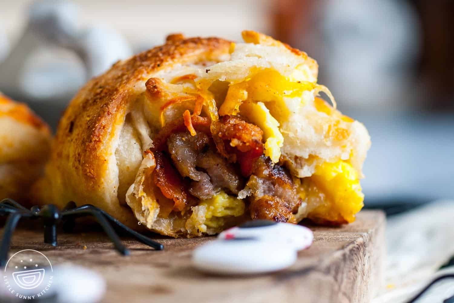 Close-up of a Halloween breakfast mummy cut open to reveal the filling of scrambled eggs, sausage, bacon, and melted cheese inside golden baked dough, with a plastic spider decoration nearby.