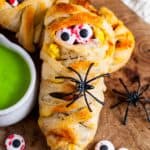 Close-up of a Halloween breakfast mummy made with pizza dough wrapped around eggs, sausage, and bacon, decorated with candy eyeballs and a plastic spider, served with bright green hollandaise sauce on the side.