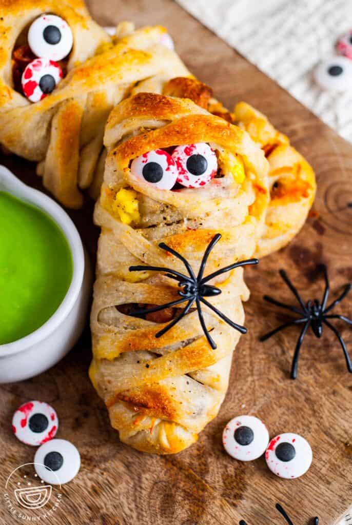 Close-up of a Halloween breakfast mummy made with pizza dough wrapped around eggs, sausage, and bacon, decorated with candy eyeballs and a plastic spider, served with bright green hollandaise sauce on the side.