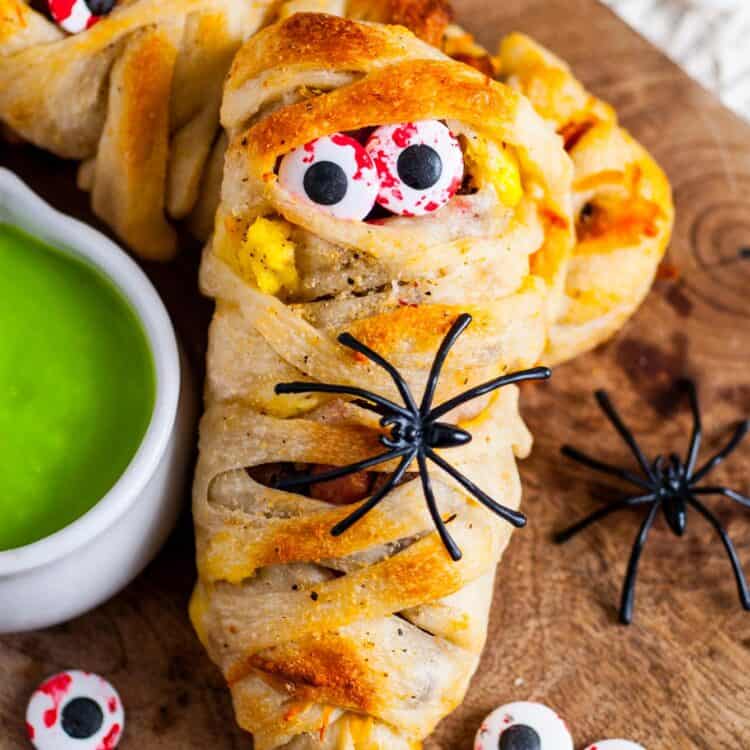 Close-up of a Halloween breakfast mummy made with pizza dough wrapped around eggs, sausage, and bacon, decorated with candy eyeballs and a plastic spider, served with bright green hollandaise sauce on the side.