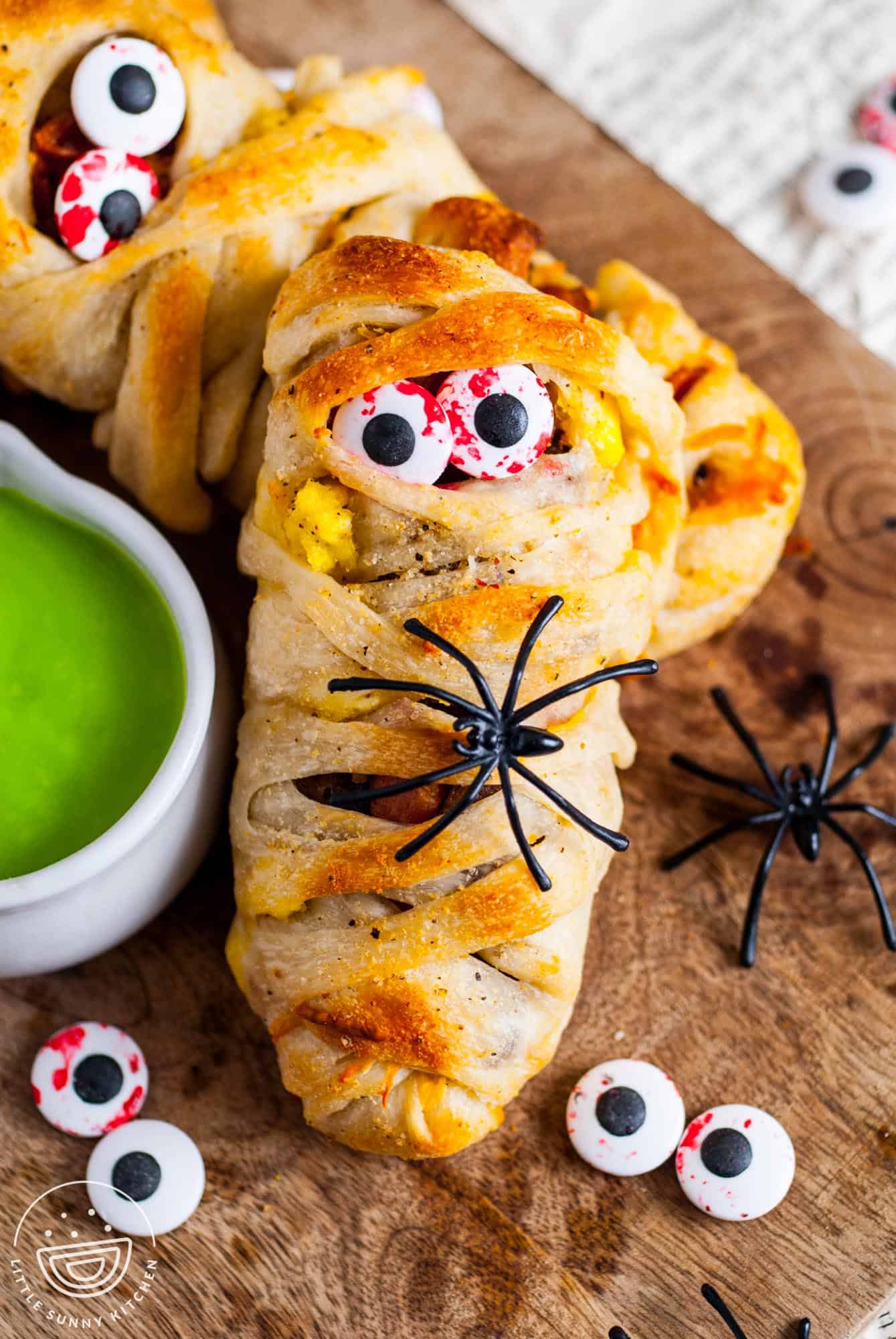 Close-up of a Halloween breakfast mummy made with pizza dough wrapped around eggs, sausage, and bacon, decorated with candy eyeballs and a plastic spider, served with bright green hollandaise sauce on the side.