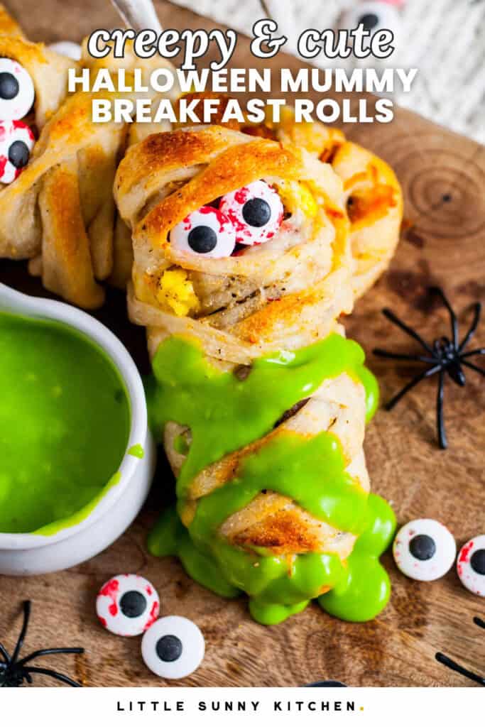 Pinterest graphic showing Halloween mummy breakfast rolls made with pizza dough wrapped around eggs, sausage, and bacon, decorated with candy eyeballs and served with bright green hollandaise sauce. Text overlay reads “Creepy & Cute Halloween Mummy Breakfast Rolls – Little Sunny Kitchen.”