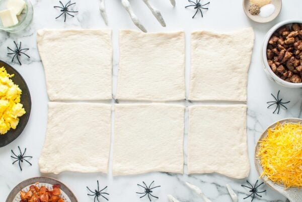 Six rectangles of pizza dough arranged on a marble surface, ready to be filled for Halloween breakfast mummies.