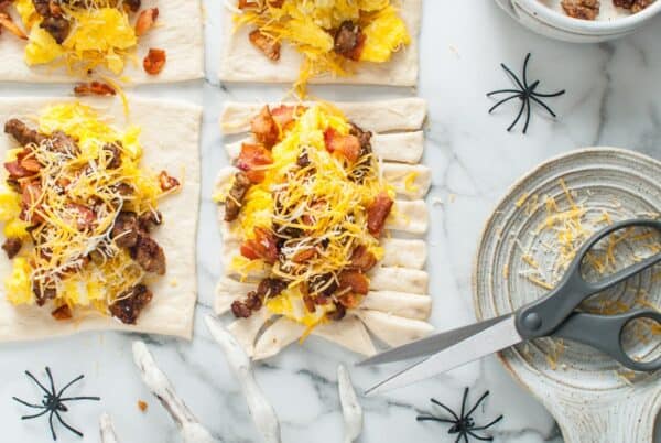 Kitchen scissors cutting fringe strips into the sides of the dough rectangles filled with eggs, sausage, bacon, and cheese.