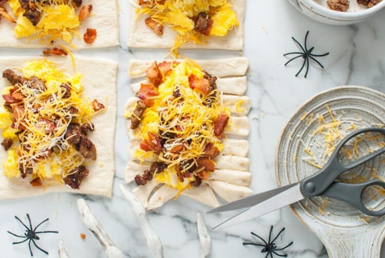 Kitchen scissors cutting fringe strips into the sides of the dough rectangles filled with eggs, sausage, bacon, and cheese.
