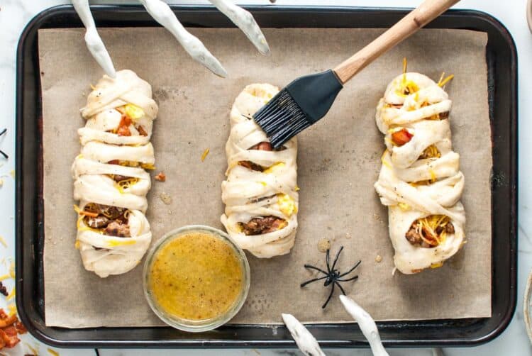 Unbaked mummies on a tray being brushed with garlic butter before going into the oven.