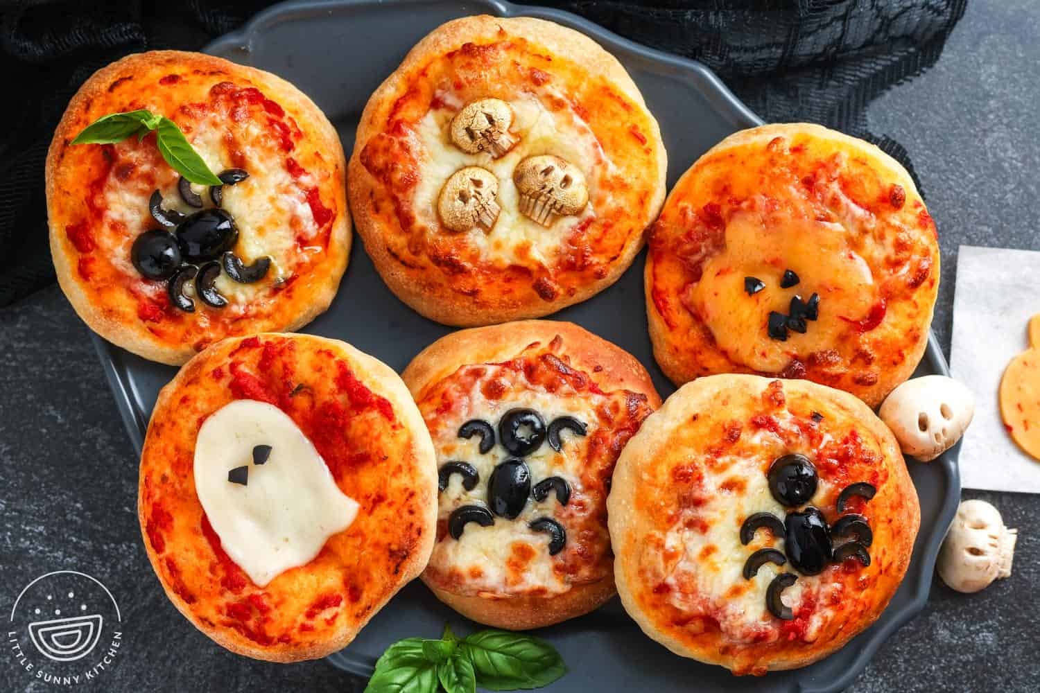 Six mini Halloween pizzas on a dark tray, each decorated with spooky designs like black olive spiders, a mozzarella ghost, a pumpkin face made of cheese, and skull shapes.