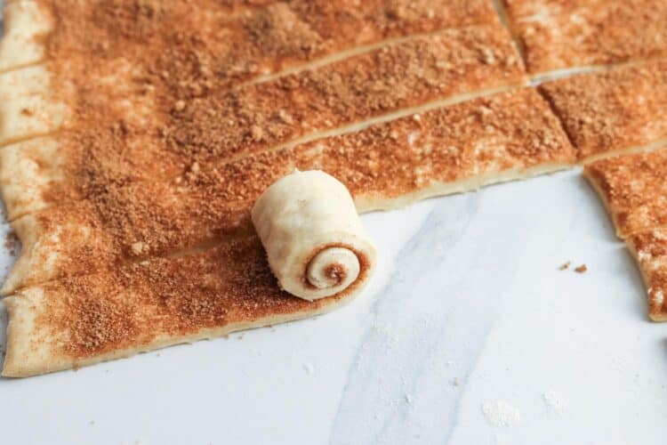 dough topped with cinnamon sugar, rolled up into mini cinnamon rolls on a marble counter.