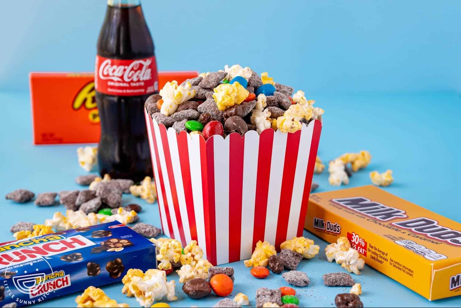 A striped popcorn box filled with movie theatre puppy chow mix made of popcorn, chocolate-coated cereal, and colorful candies, surrounded by candy boxes and a Coke bottle on a blue background.