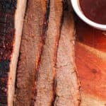 sliced slow cooked beef brisket on a wooden cutting board next to a small bowl of barbecue sauce.