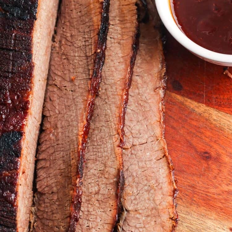 sliced slow cooked beef brisket on a wooden cutting board next to a small bowl of barbecue sauce.