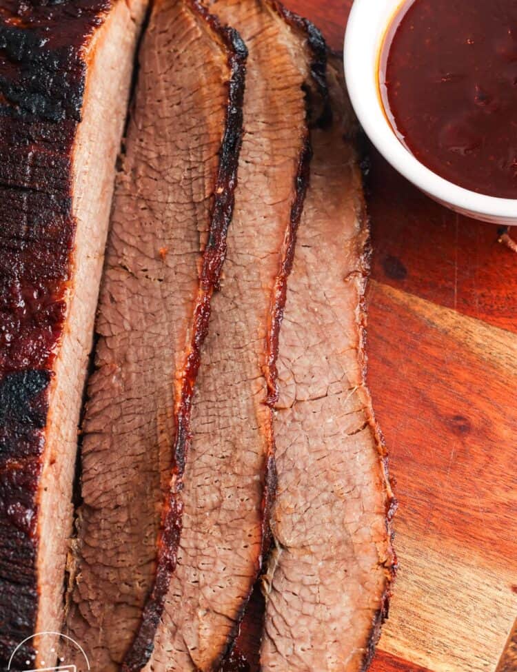 sliced slow cooked beef brisket on a wooden cutting board next to a small bowl of barbecue sauce.
