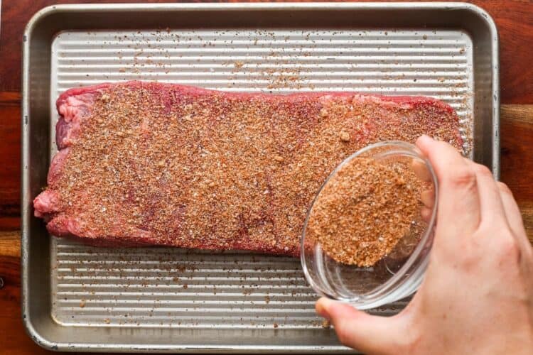 a hand sprinkling spice rub over a large beef brisket that is on a metal sheet pan.