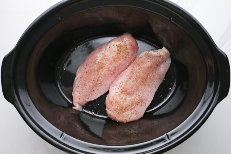 two chicken breasts at the bottom of a slow cooker, seasoned with herbs.