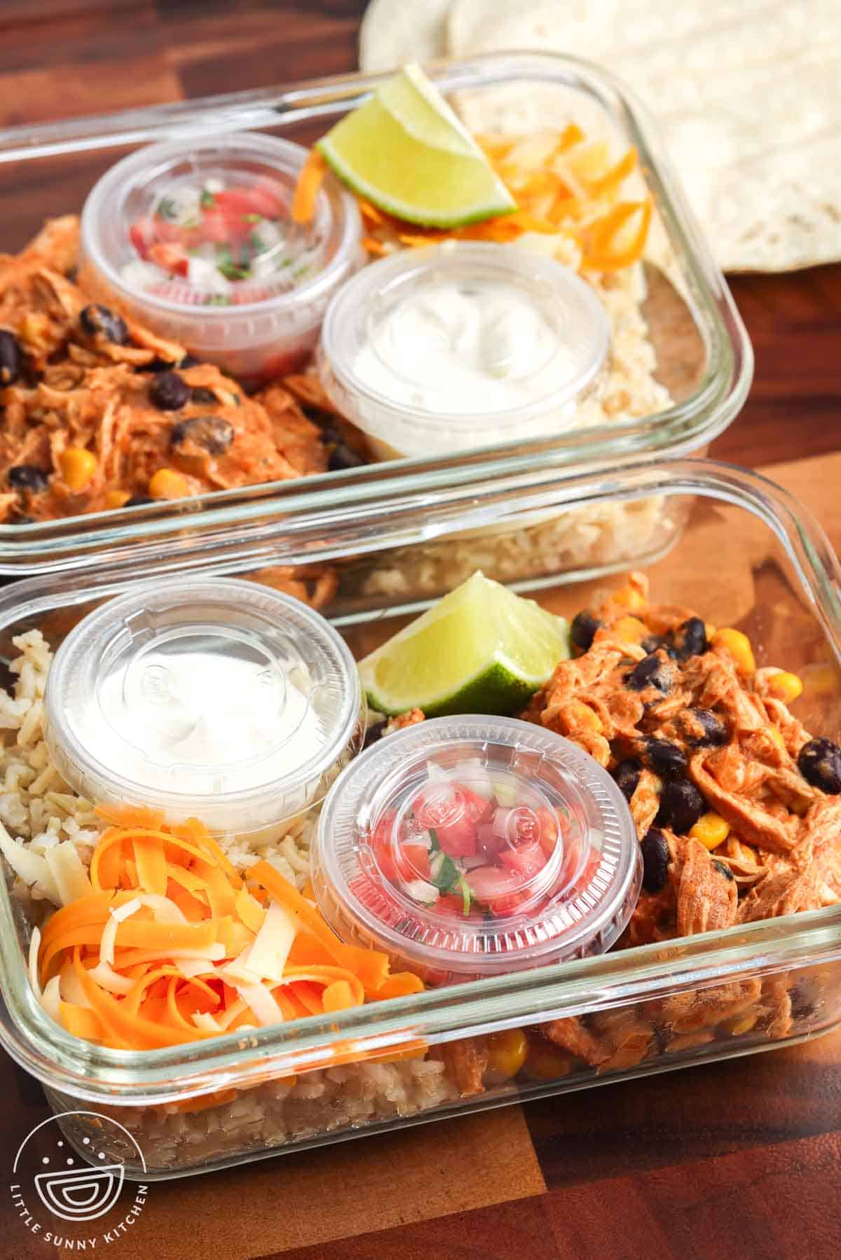 Two glass meal prep containers filled with slow cooker chicken enchilada bowls with rice, shredded chicken, black beans, corn, shredded cheese, sour cream, pico de gallo, and lime wedges.