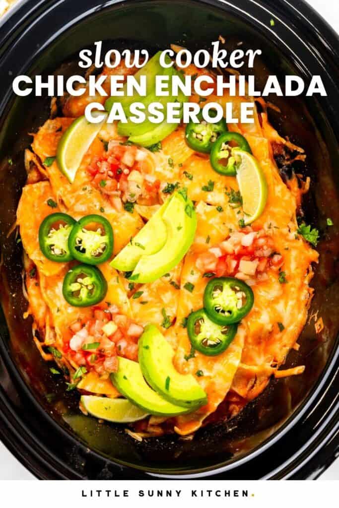 cheesy casserole in a crock top, topped with pico de gallo, avocado slices, and sliced jalapenos. Text overlay says "slow cooker chicken enchilada casserole"