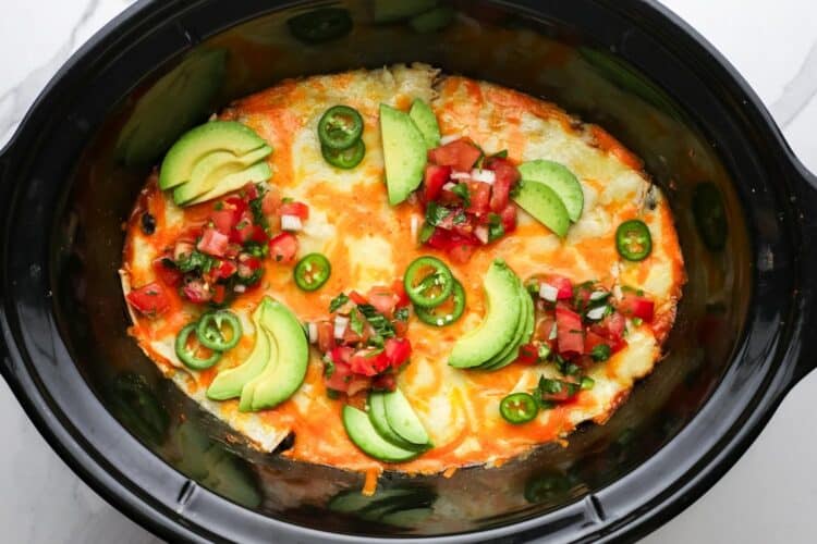 Slow cooker chicken enchilada casserole with melty cheese on top, avocado, pico de gallo, and sliced jalapeno, in a black oval crock pot.