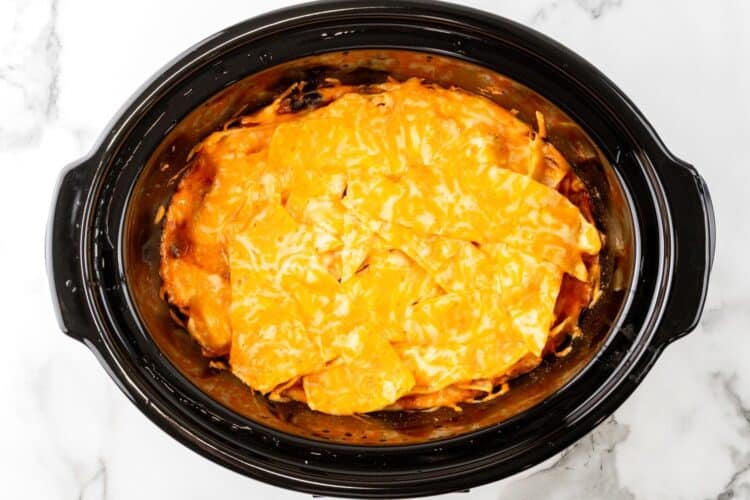 slow cooker chicken enchilada casserole with melty cheese on top, in a black oval crock pot.