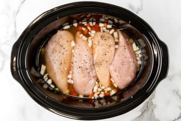 chicken breasts in a slow cooker surrounded by broth, chopped carrots, and onions.