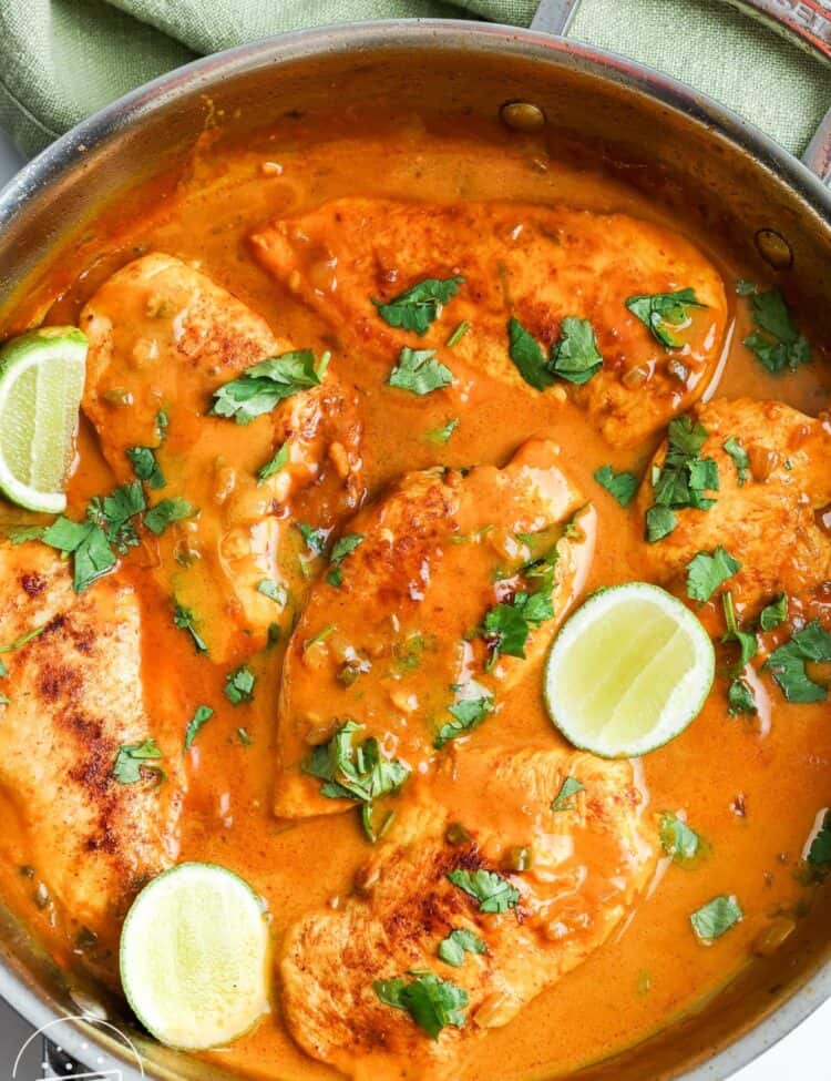a skillet of cooked chicken with coconut lime sauce, and slices of fresh lime and cilantro for garnish.