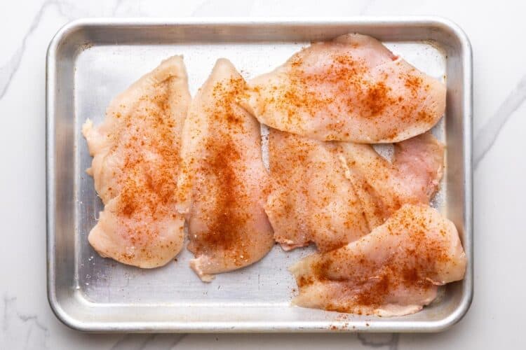 five chicken cutlets on a metal tray, seasoned with salt and paprika