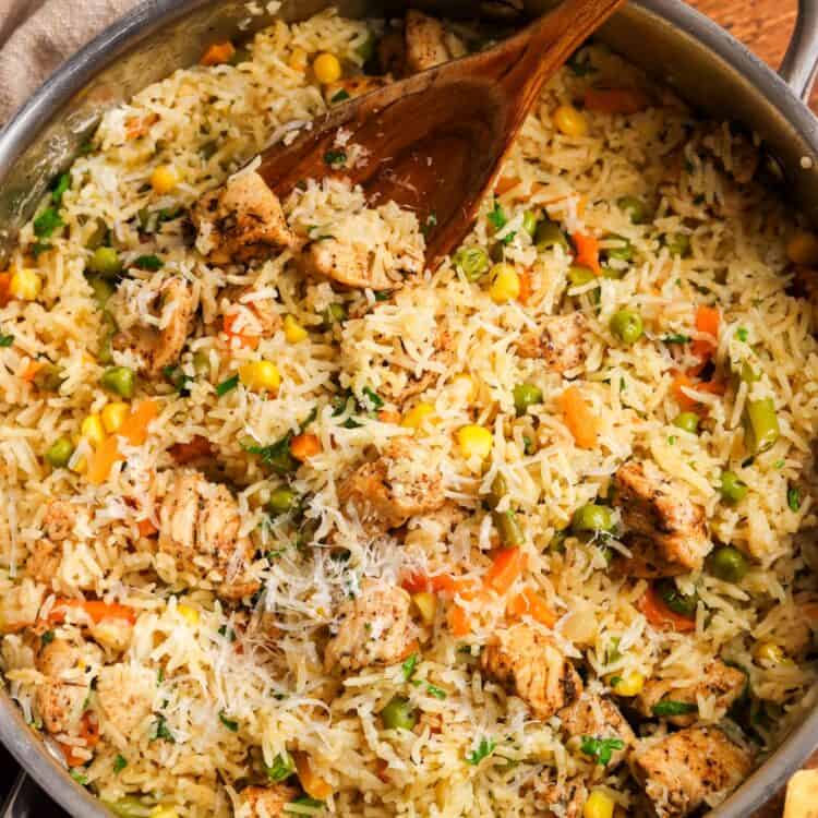 a large stainless steel pot filled with fluffy rice with mixed veggies and chicken, topped with parmesan cheese. The one pot chicken and rice is being served with a large wooden spoon.