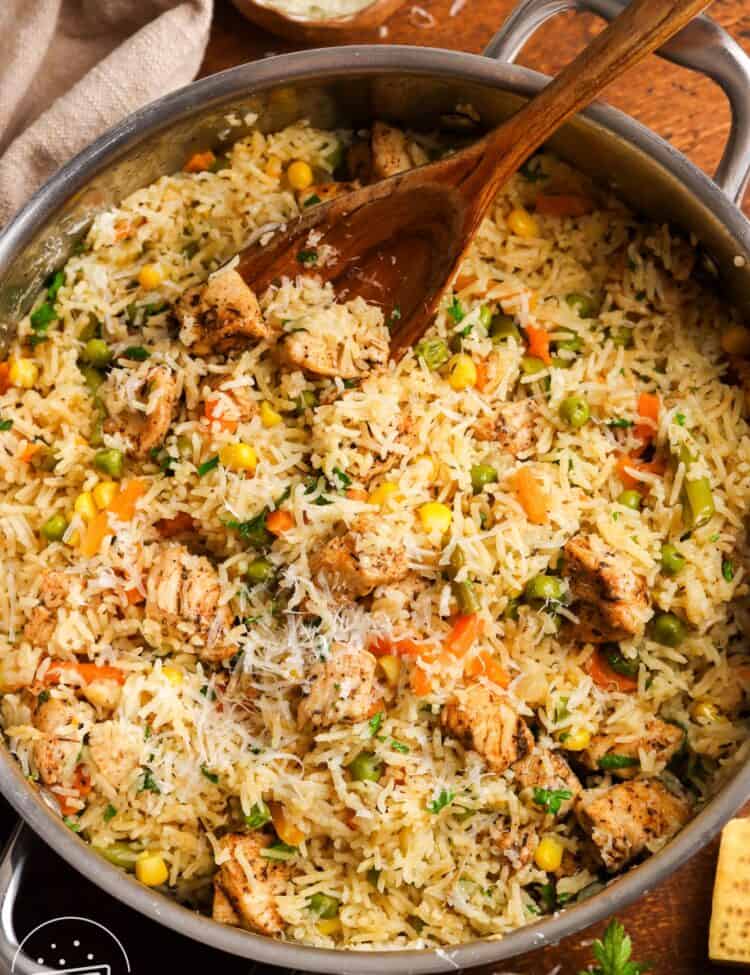 a large stainless steel pot filled with fluffy rice with mixed veggies and chicken, topped with parmesan cheese. The one pot chicken and rice is being served with a large wooden spoon.