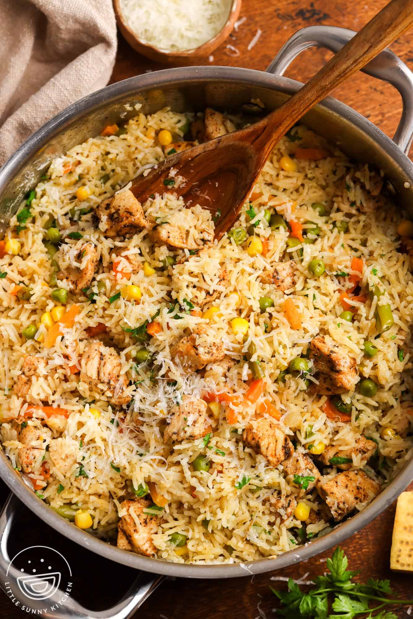 a large stainless steel pot filled with fluffy rice with mixed veggies and chicken, topped with parmesan cheese. The one pot chicken and rice is being served with a large wooden spoon.