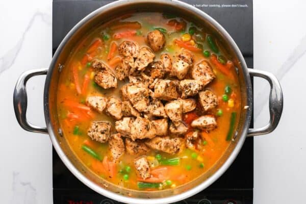 cooked chicken breast pieces added to a pot of rice cooking with broth and frozen veggies.