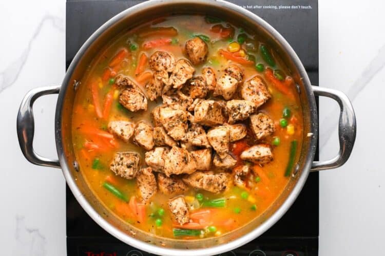 cooked chicken breast pieces added to a pot of rice cooking with broth and frozen veggies.