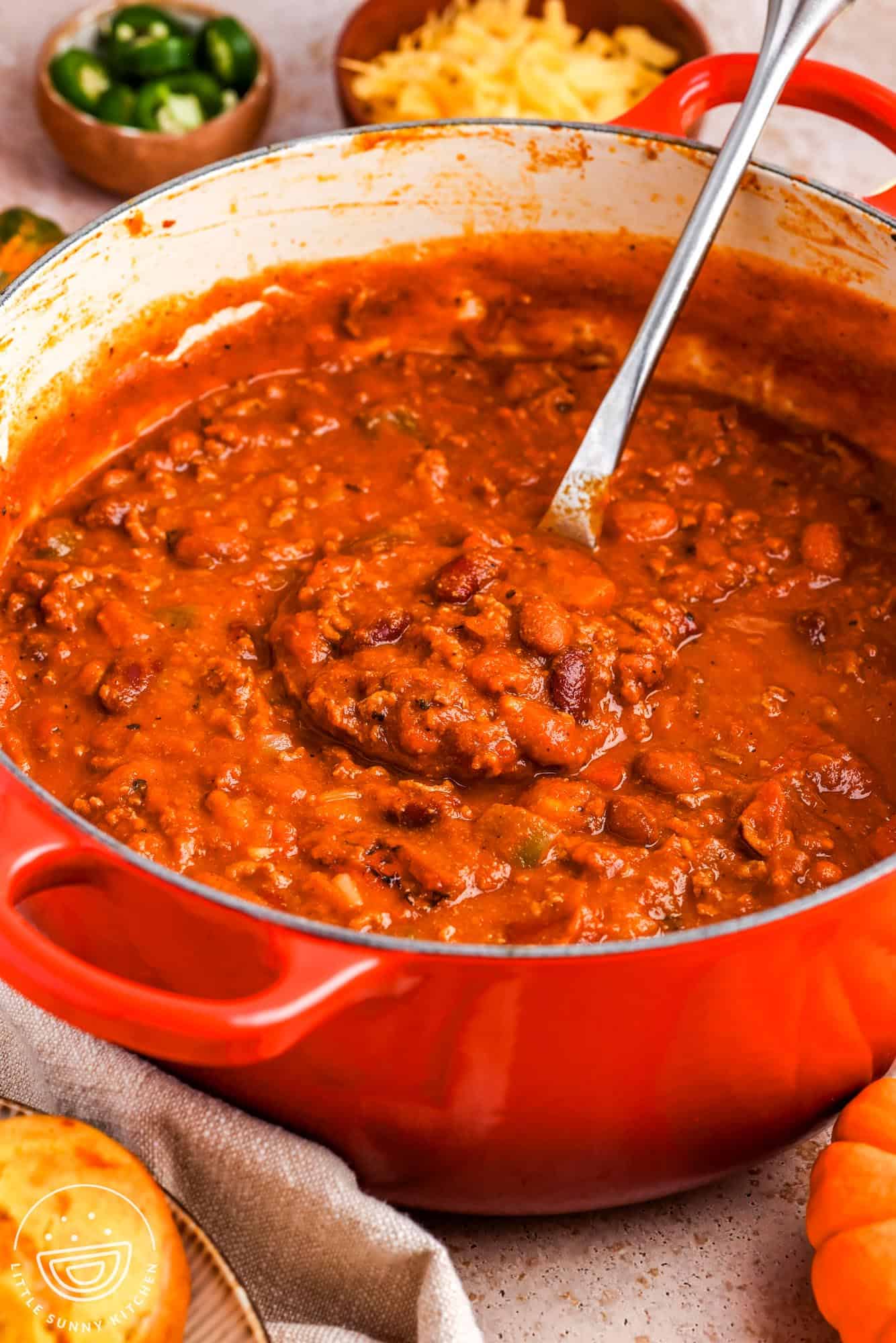 a red dutch oven filled with thick, deep red beef, bean, and pumpkin chili. In the background are bowls with garnishes, including peppers and shredded cheese.