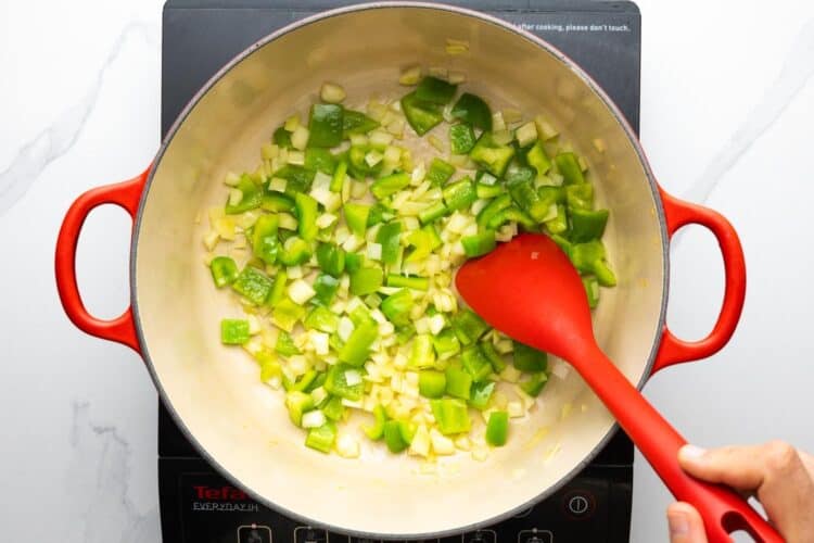 green bell peppers and diced onion sauteed in a dutch oven, stirred with a red spatula.