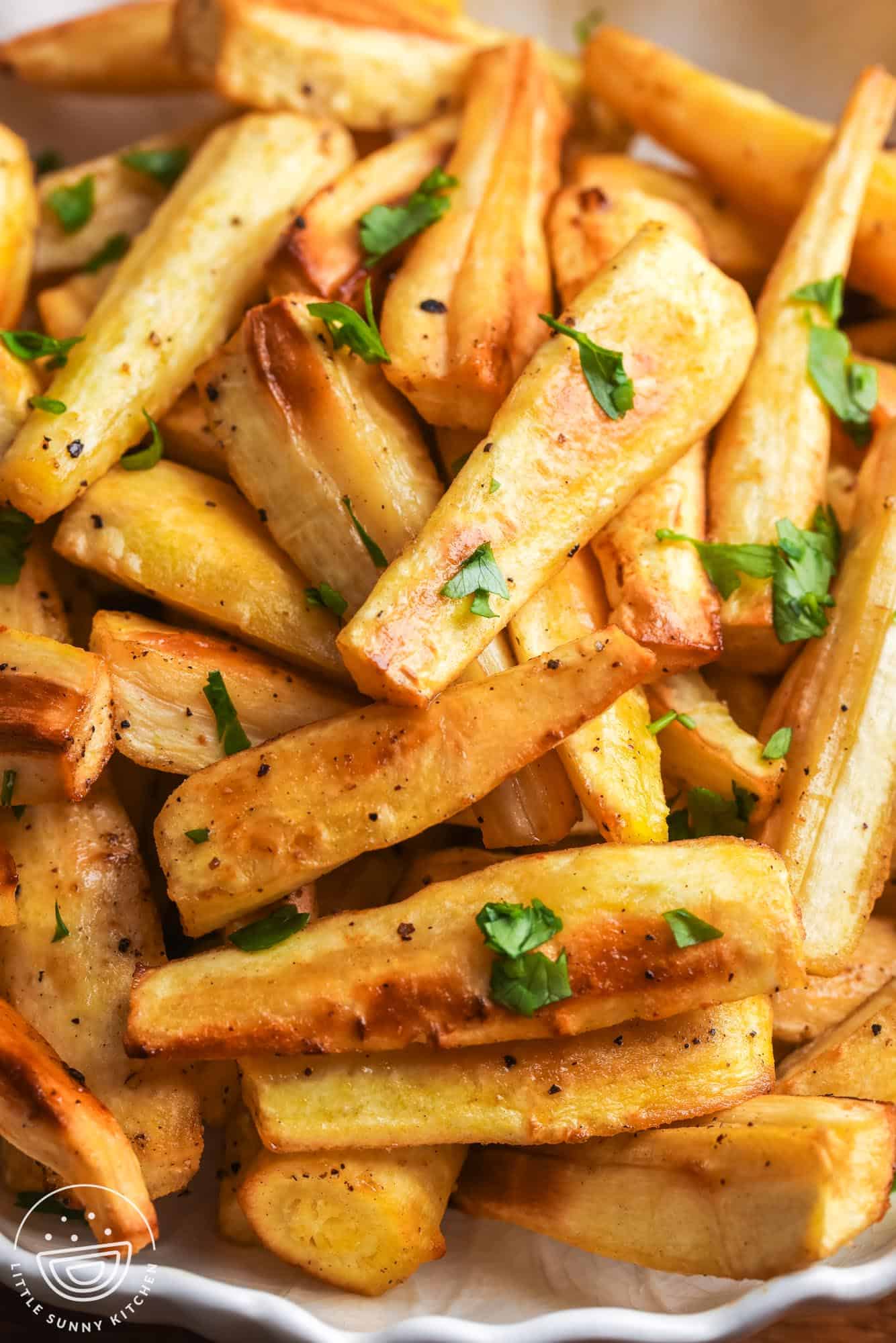 Close-up of roasted parsnips with crispy edges and chopped parsley in a white serving bowl.