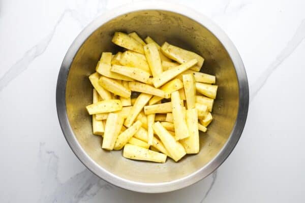 Peeled and cut raw parsnip batons in a mixing bowl tossed with olive oil, honey, salt, and pepper.
