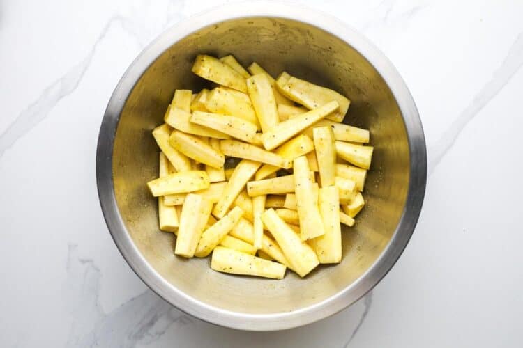 Peeled and cut raw parsnip batons in a mixing bowl tossed with olive oil, honey, salt, and pepper.