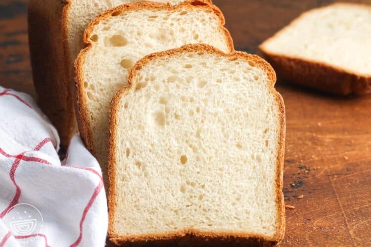 slices of homemade sandwich bread on a wooden cutting board, next to a white and red kitchen towel.