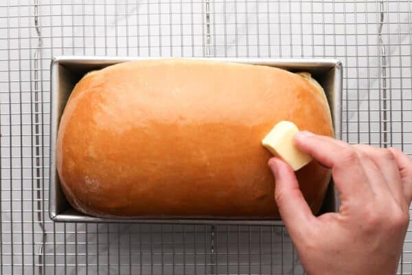 a hand rubbing a pat of butter over a warm baked loaf of homemade sandwich bread.