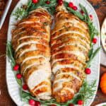 sliced meat from a bone-in turkey breast on a white platter, garnished with fresh green herbs and cranberries.